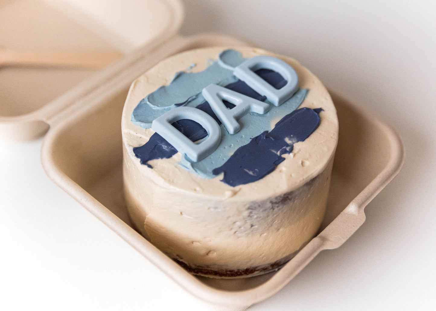 Small round cake with 'DAD' on top in a beige box on a white background